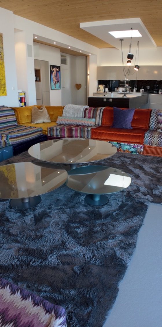 Modern Wohnzimmer mit bunten Sofas, Glastischen und einem Teppich. Kochnische im Hintergrund.