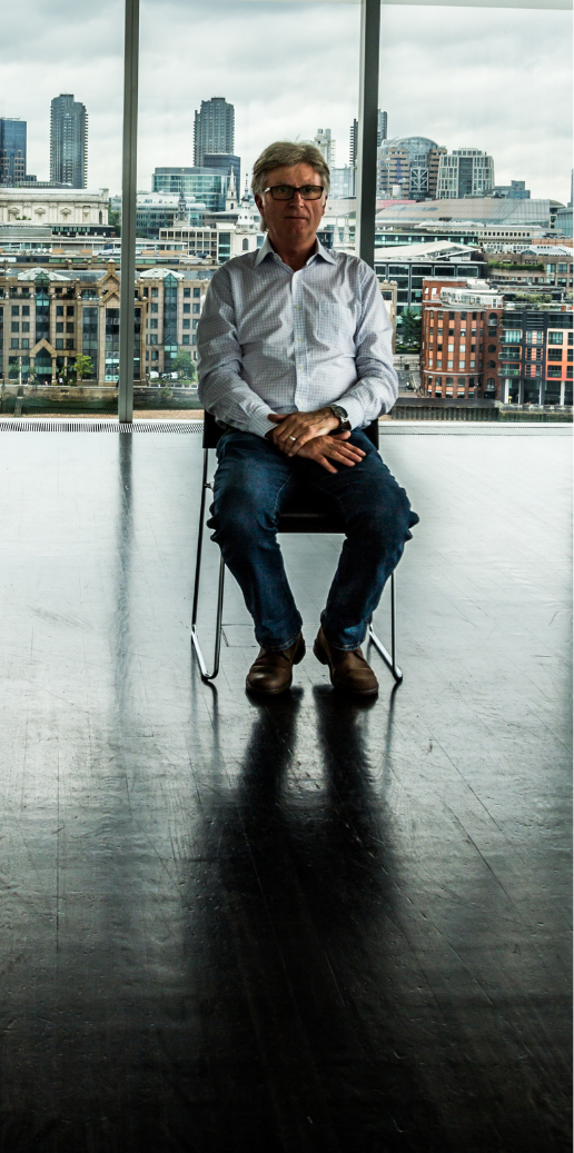 Mann in Hemd sitzt auf einem Stuhl vor einem großen Fenster mit Stadtblick.